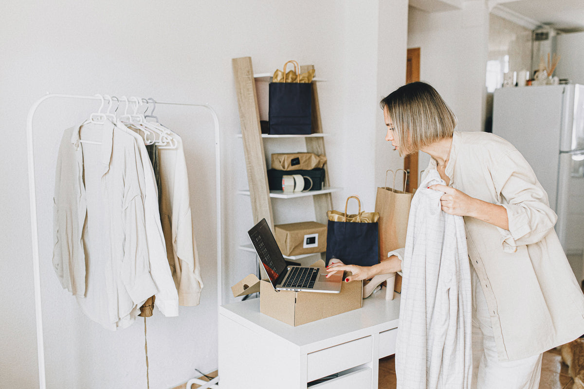 A woman in a light beige linen outfit stands in a bright, modern room, holding a white towel while looking at a laptop on a white desk. To her left is a white clothing rack with neutral-toned garments, and behind her are shelves with bags and boxes.